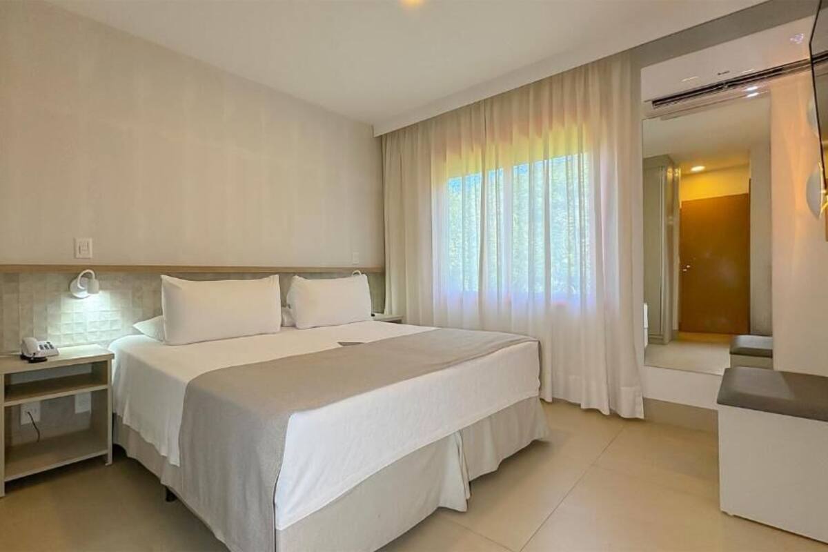A hotel room with a bed, bedside table, lamp, and a view of the outdoors through the window.