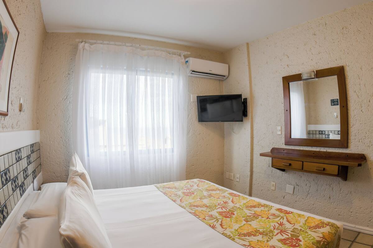 A hotel room with a bed, a TV mounted on the wall, a window with curtains, and a small wooden shelf.