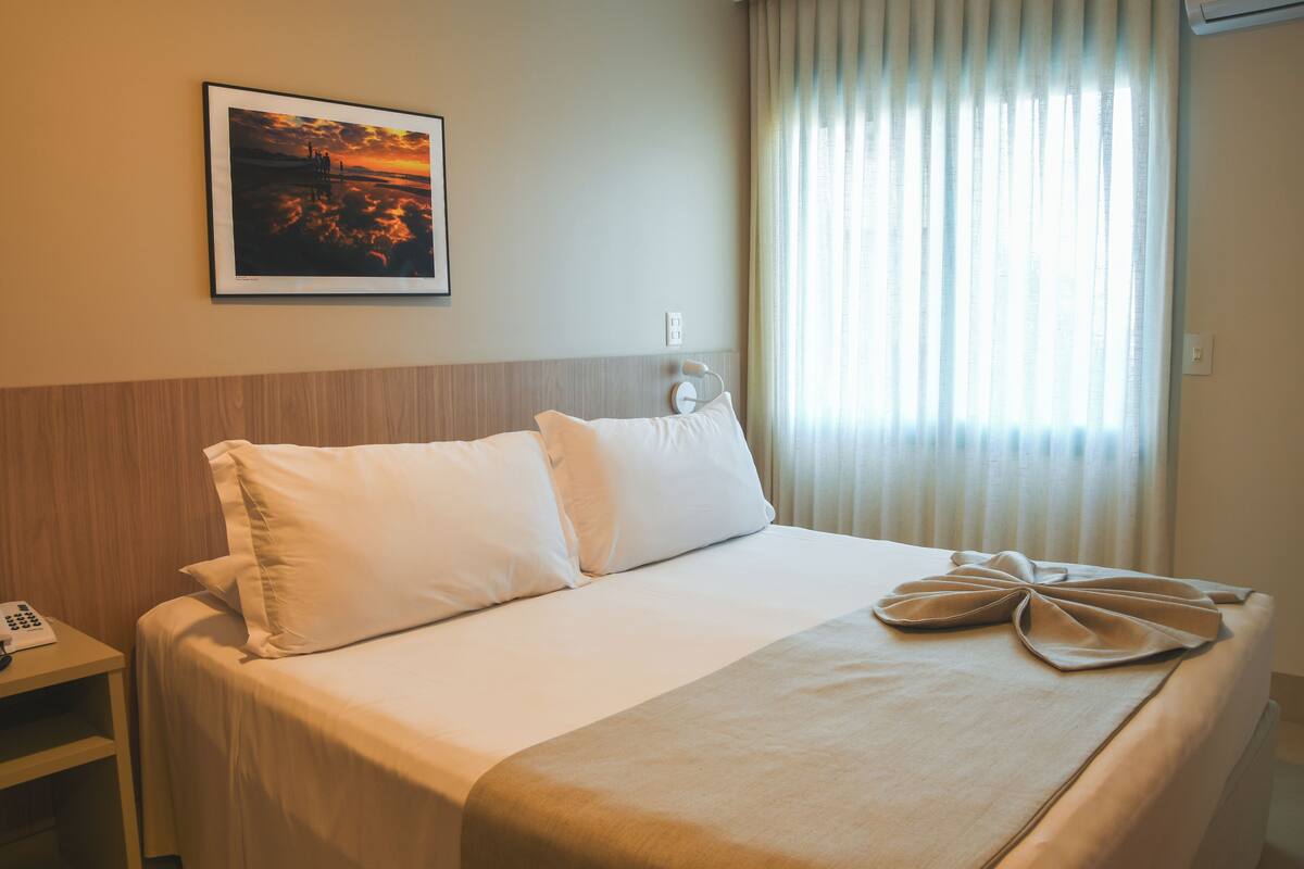 A hotel room with a bed, a bedside table with a phone, a framed picture on the wall, and a window with curtains.