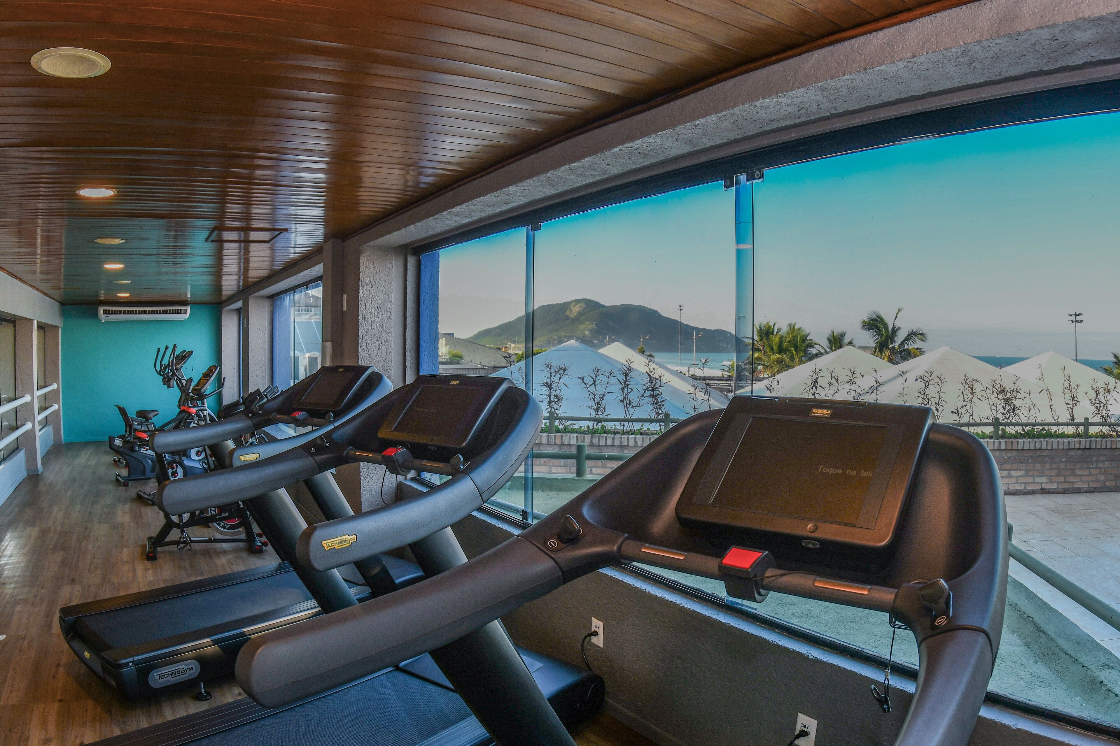 Un gimnasio con cintas de correr y bicicletas estáticas, amplias ventanas con vistas a montañas y palmeras.