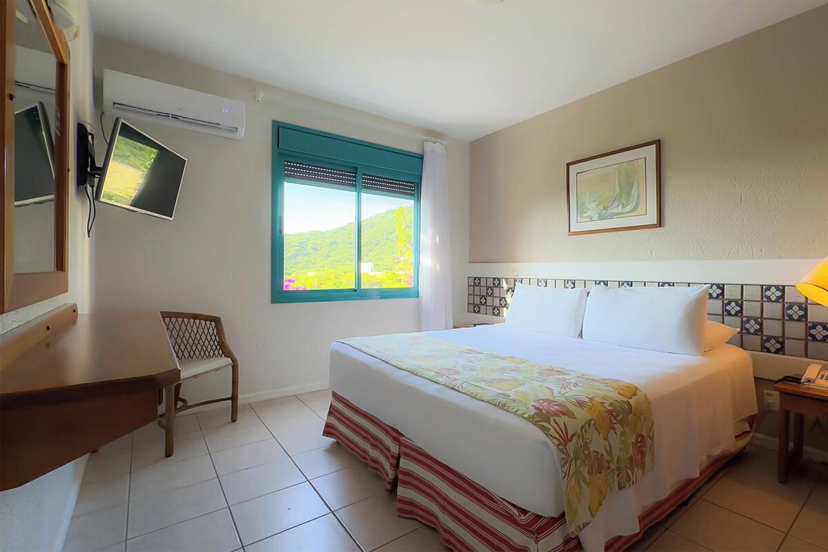 A hotel room with a bed, a desk, a chair, a window with a view, an air conditioner, a lamp, and a telephone.