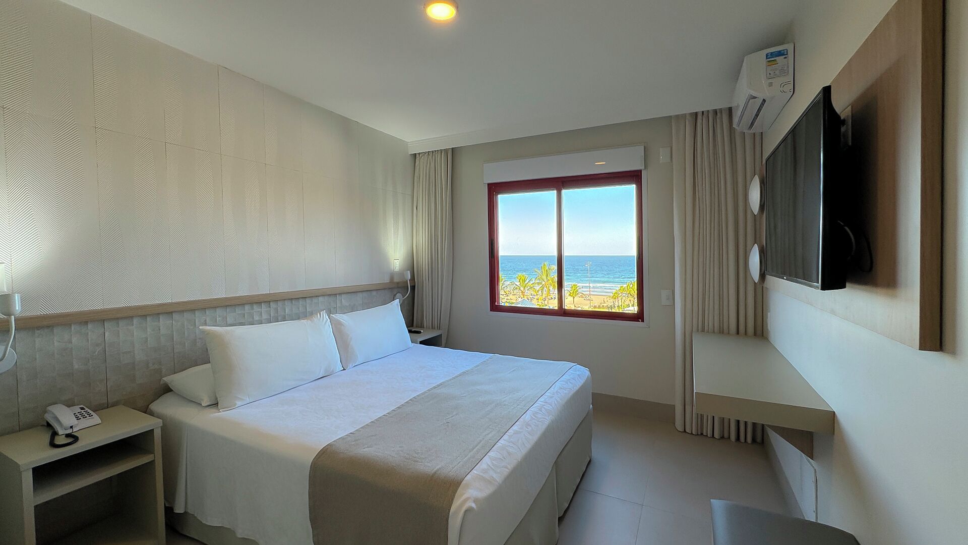 A hotel room with a bed, a TV, a phone on the nightstand, and a window with a view of the beach and ocean.