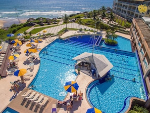 A large outdoor swimming pool with multiple lanes, surrounded by lounge chairs and umbrellas, and a view of the ocean. - Costao do Santinho Resort - All Inclusive (Florianópolis)