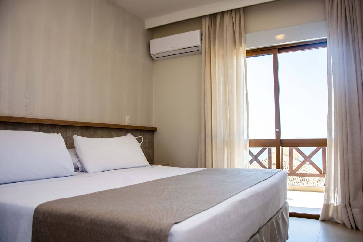 A hotel room with a bed, an air conditioning unit, a balcony with a view, and curtains.