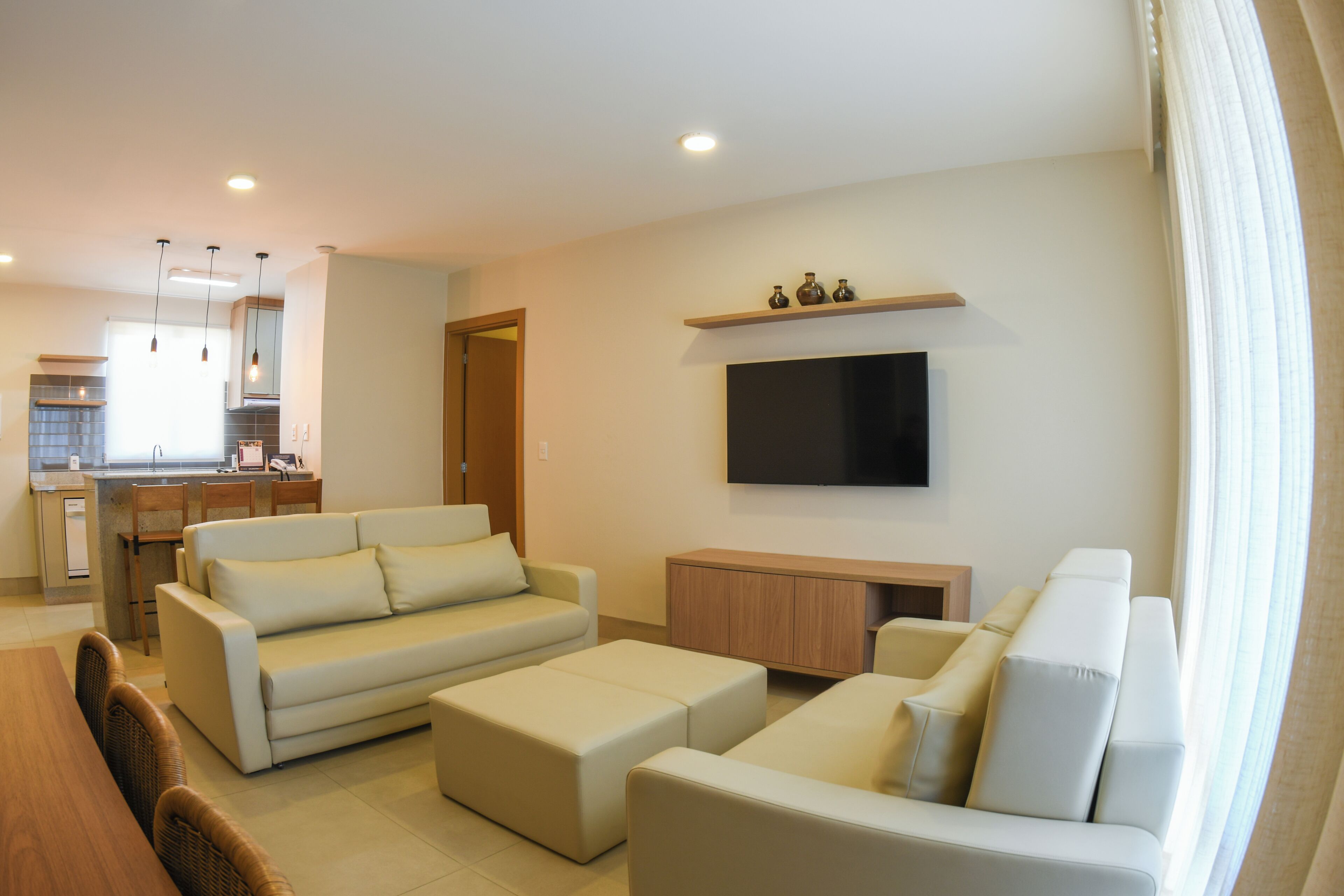 A modern living room with a sofa set, a TV mounted on the wall, and a kitchen in the background.