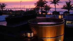 Une terrasse en bois avec un jacuzzi et des plantes en pot, donnant sur une plage au coucher du soleil.