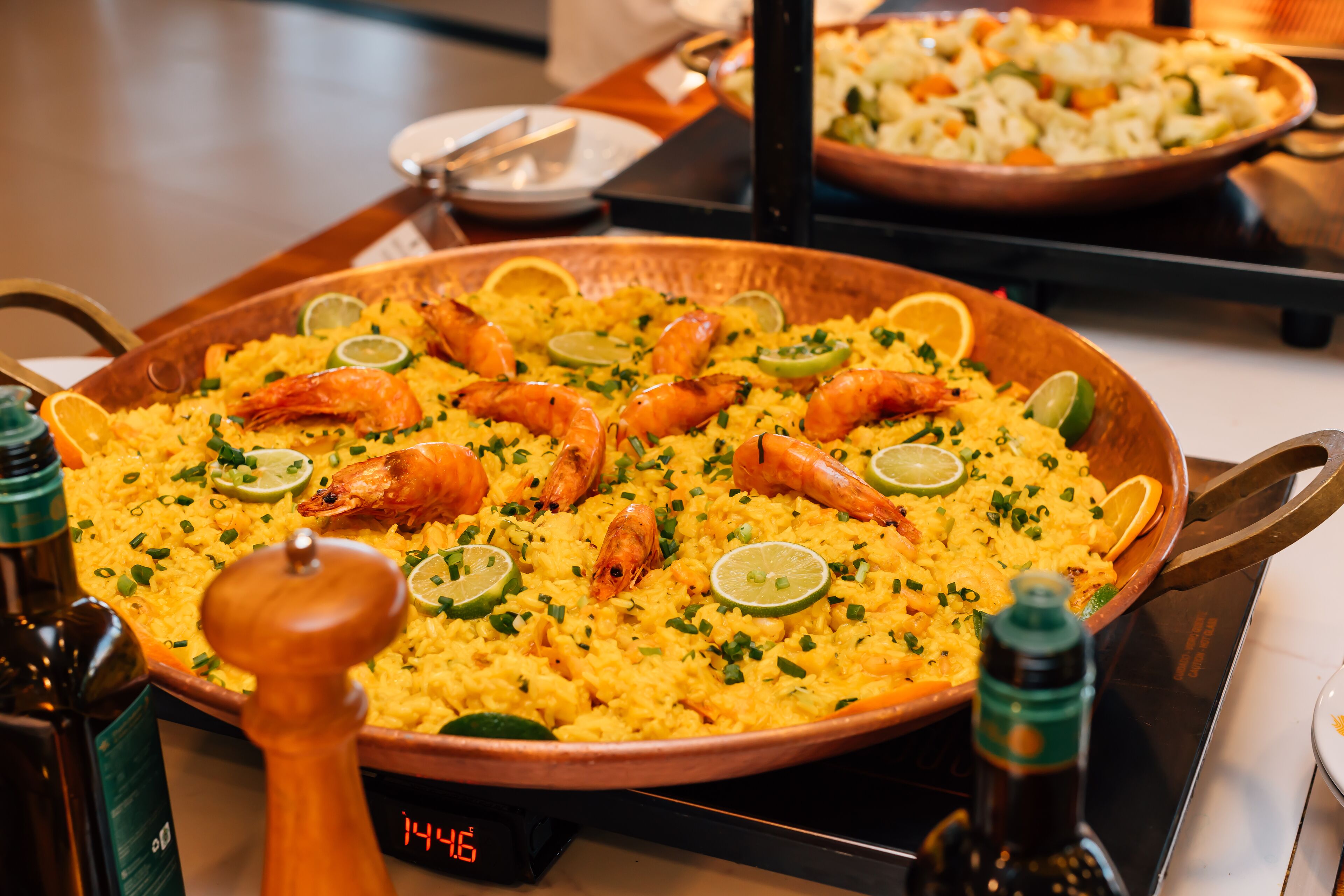 Una olla de cobre grande llena de arroz amarillo, adornada con camarones y rodajas de lima, sobre una estufa.