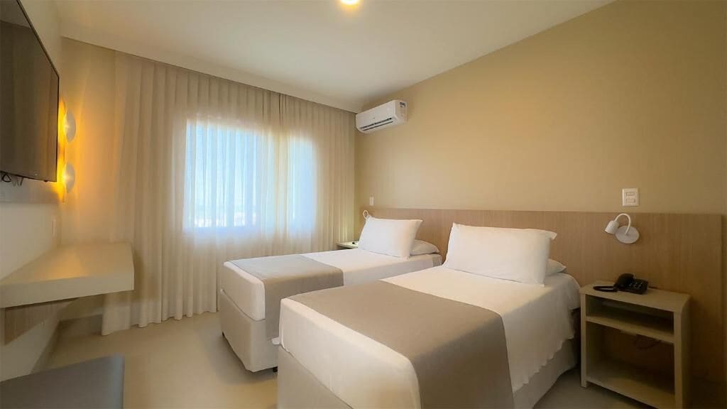 Quarto de hotel com duas camas, televisão, ar condicionado e mesa de cabeceira com telefone.