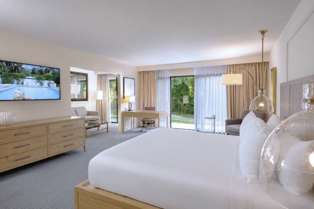 A hotel room with a large bed, a television mounted on the wall, a desk, and a view of the outdoors through the windows.