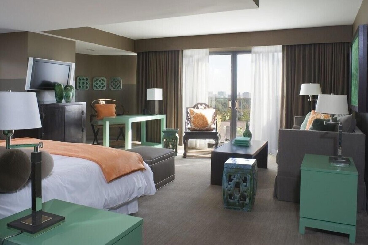 A hotel room with a bed, two armchairs, a sofa, a desk, a television, and a balcony with curtains.