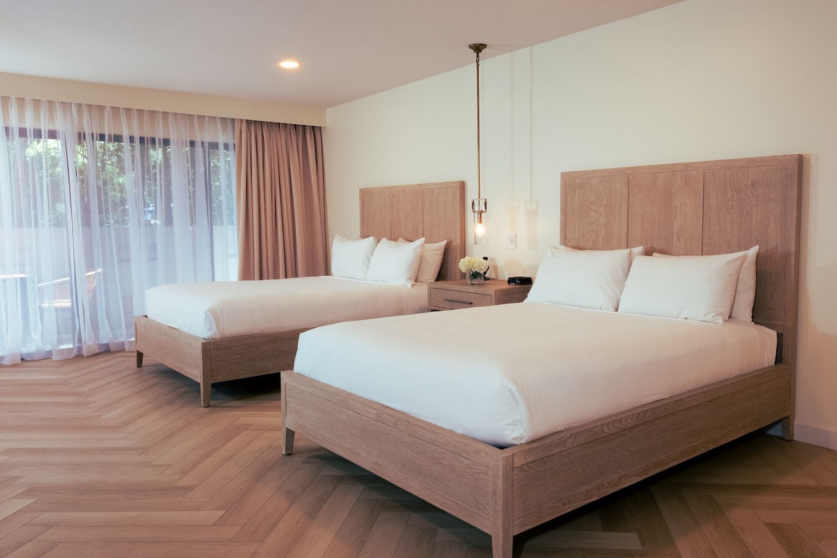 A hotel room with two beds, wooden headboards, a nightstand, a lamp, and sheer curtains.