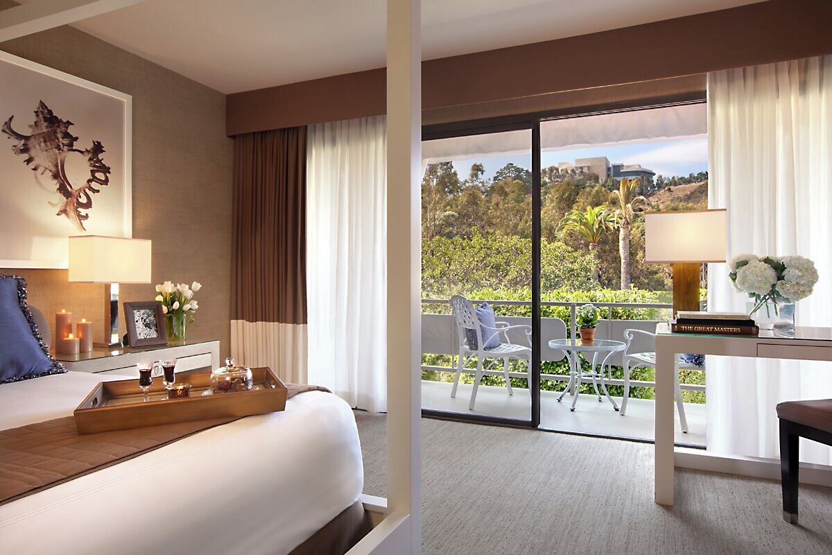 A hotel room with a bed, a bedside table with a lamp, a tray with glasses, and a view of the outdoors through a sliding door.