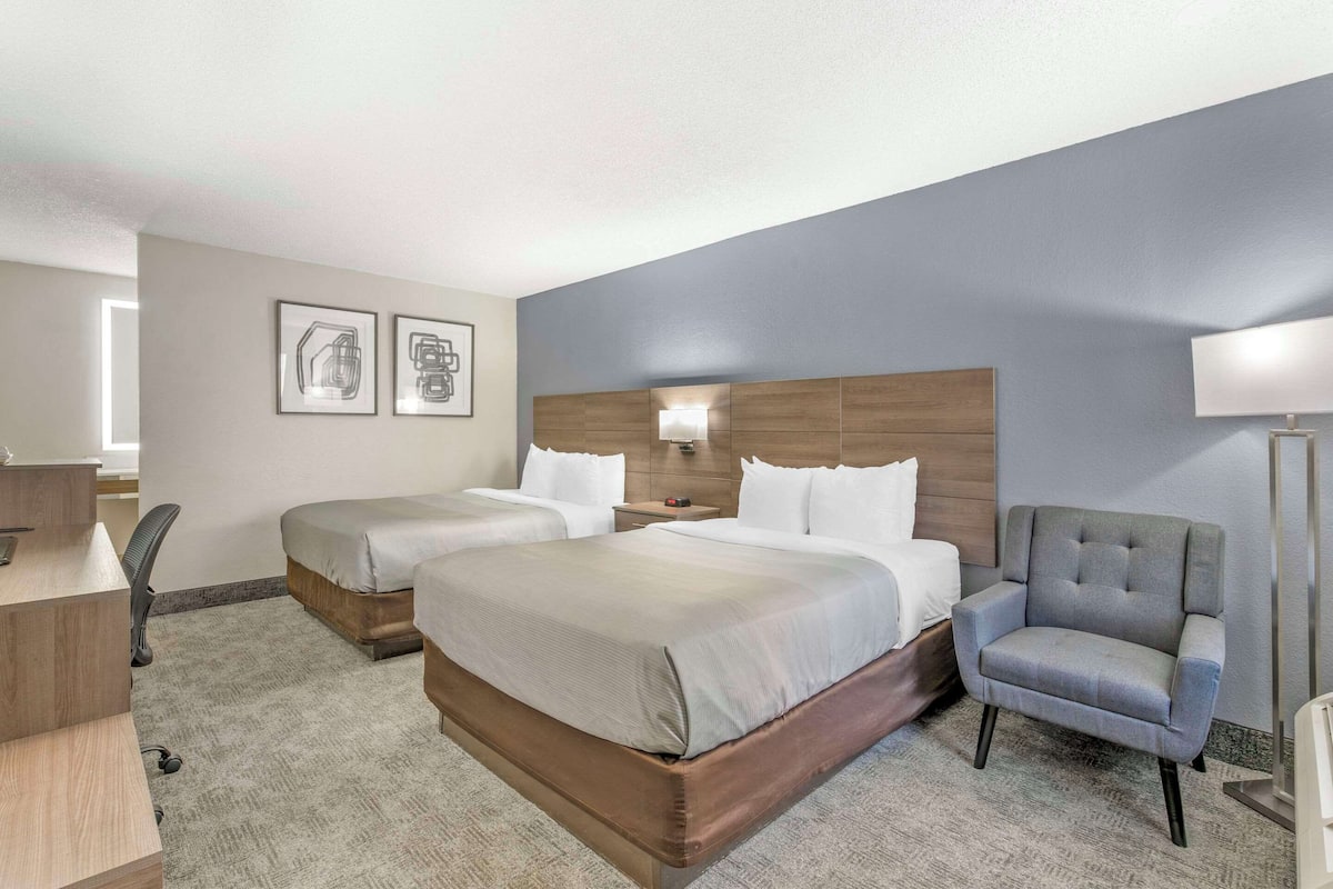 A hotel room with two beds, a desk, a chair, and a lamp.