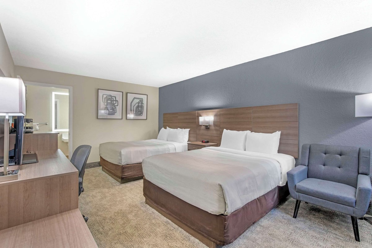A hotel room with a large bed, two smaller beds, a desk with a chair, a grey armchair, and a wall-mounted TV.