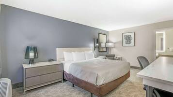 A hotel room with a bed, bedside table, lamp, chair, and a desk with a chair.