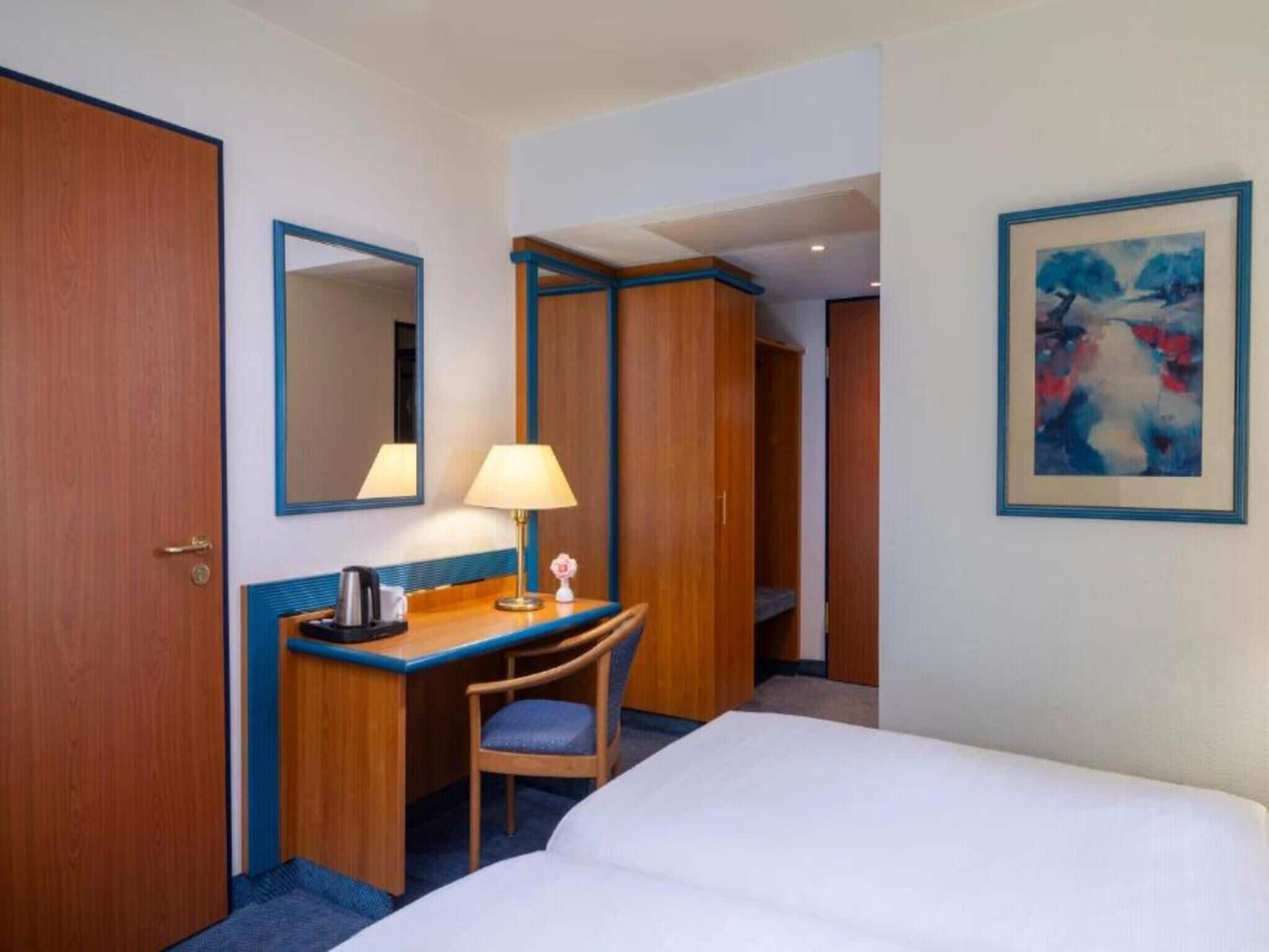 Habitación de hotel con cama, escritorio con silla, lámpara, espejo y un cuadro en la pared.