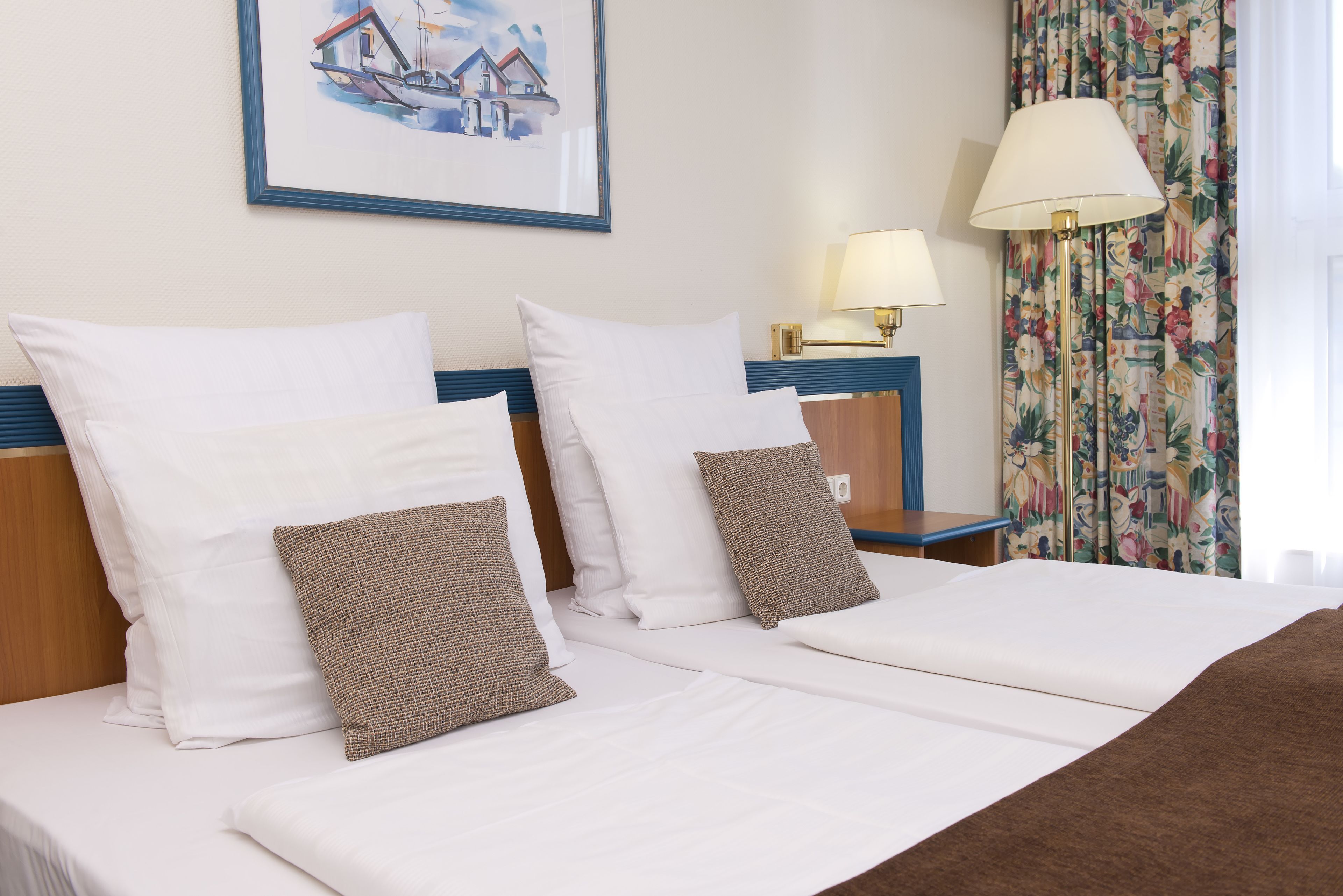 Une chambre d’hôtel avec deux lits, du linge de lit blanc et un tableau représentant une maison accroché au mur.