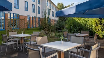 Eine Terrasse im Freien mit Tischen und Stühlen, ein Gebäude mit blauen Fenstern im Hintergrund und ein Hecke im Vordergrund.