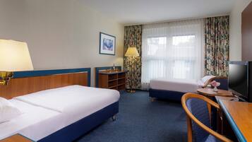 Ein Hotelzimmer mit zwei Betten, einem Schreibtisch mit Stuhl, einem Fernseher und einem Fenster mit Vorhängen.
