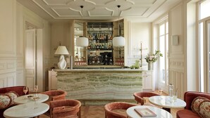 A bar area with a marble countertop, multiple shelves of bottles, and a bar counter. There are two round tables with chairs, and a sofa with floral cushions. - Le 5 Particulier (Neuilly-sur-Seine)