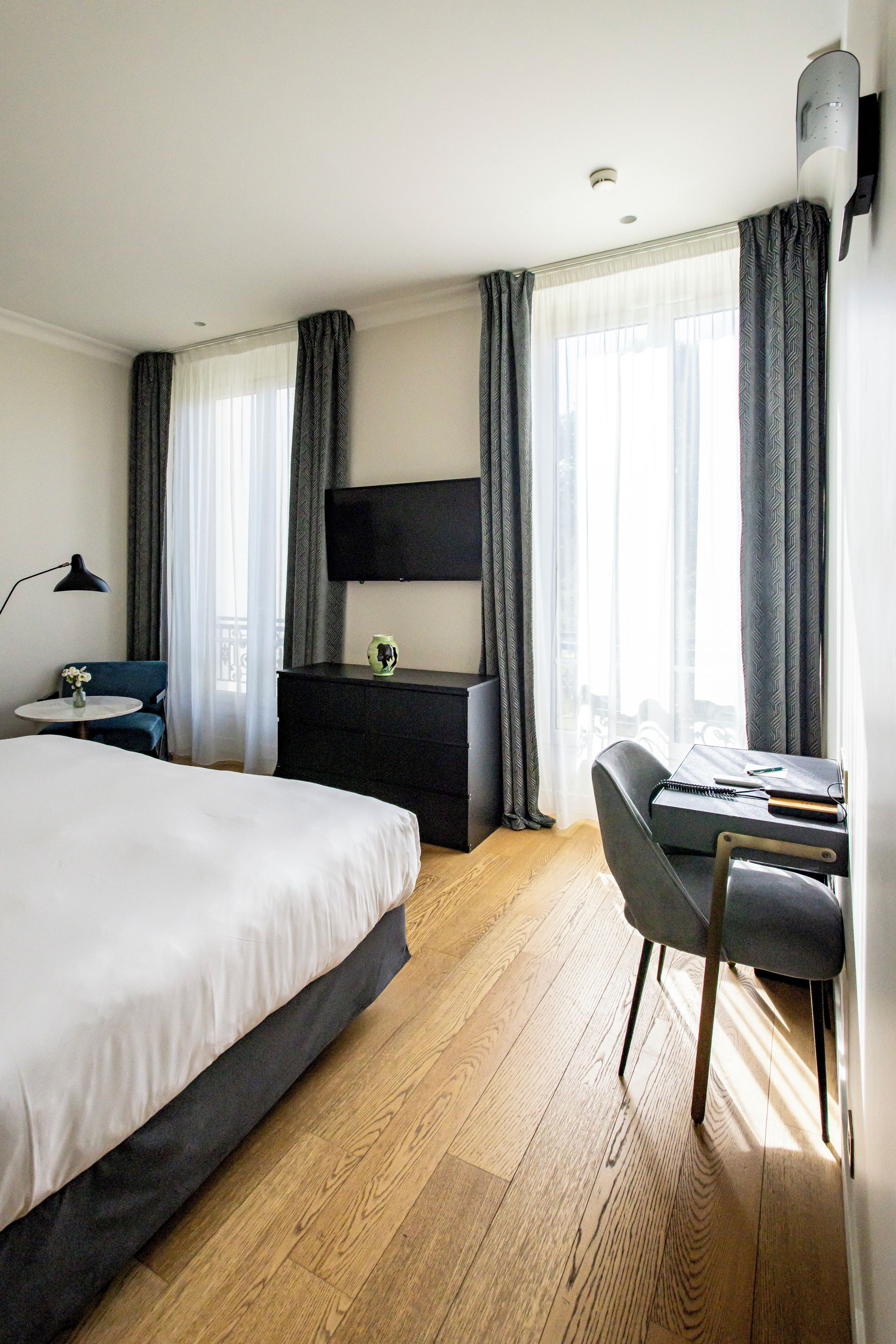 A hotel room with a large bed, a desk with a chair, a flat-screen TV mounted on the wall, and a large window with curtains.