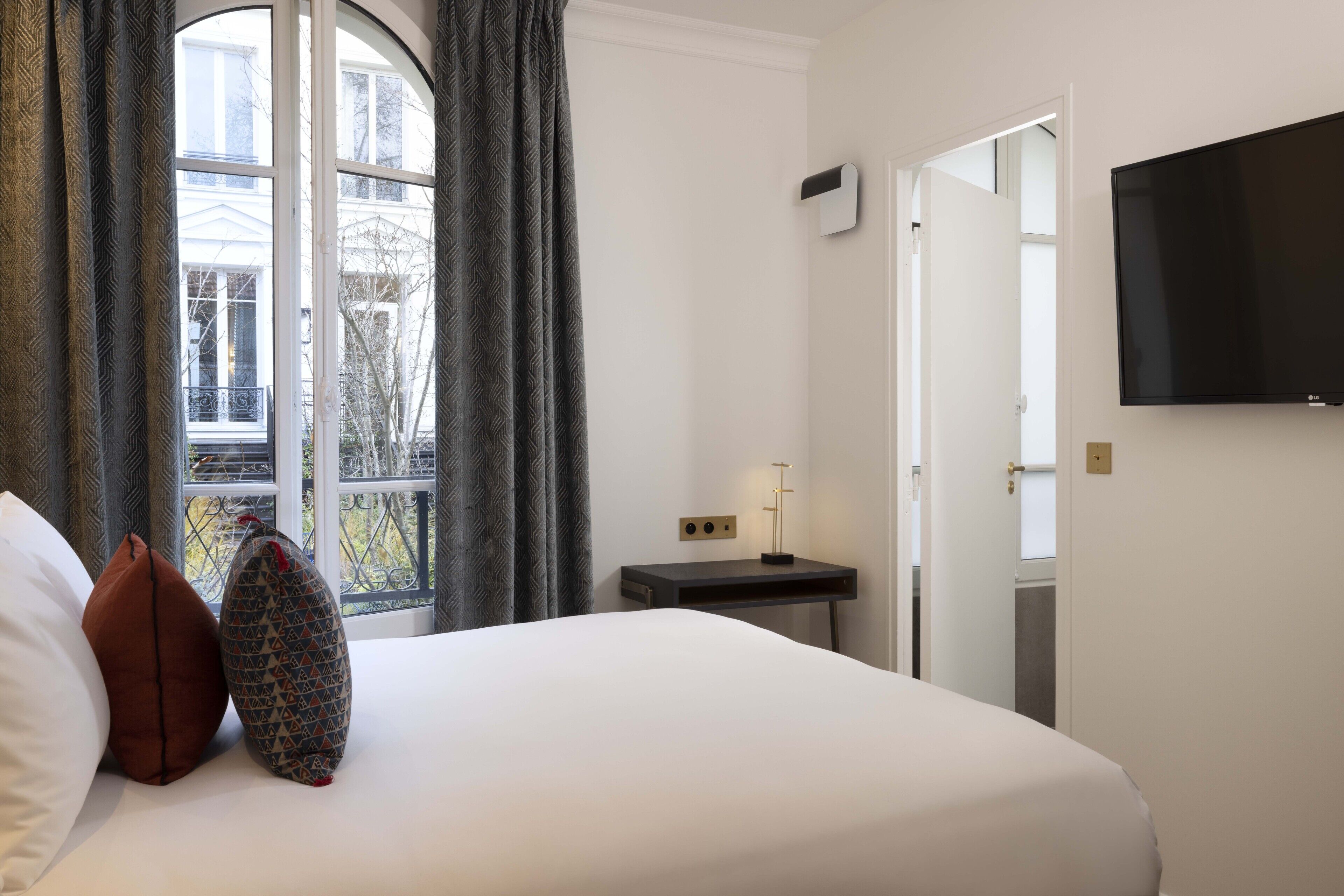 Una habitación de hotel con cama, mesita de noche, televisor de pantalla plana y ventana con cortinas.