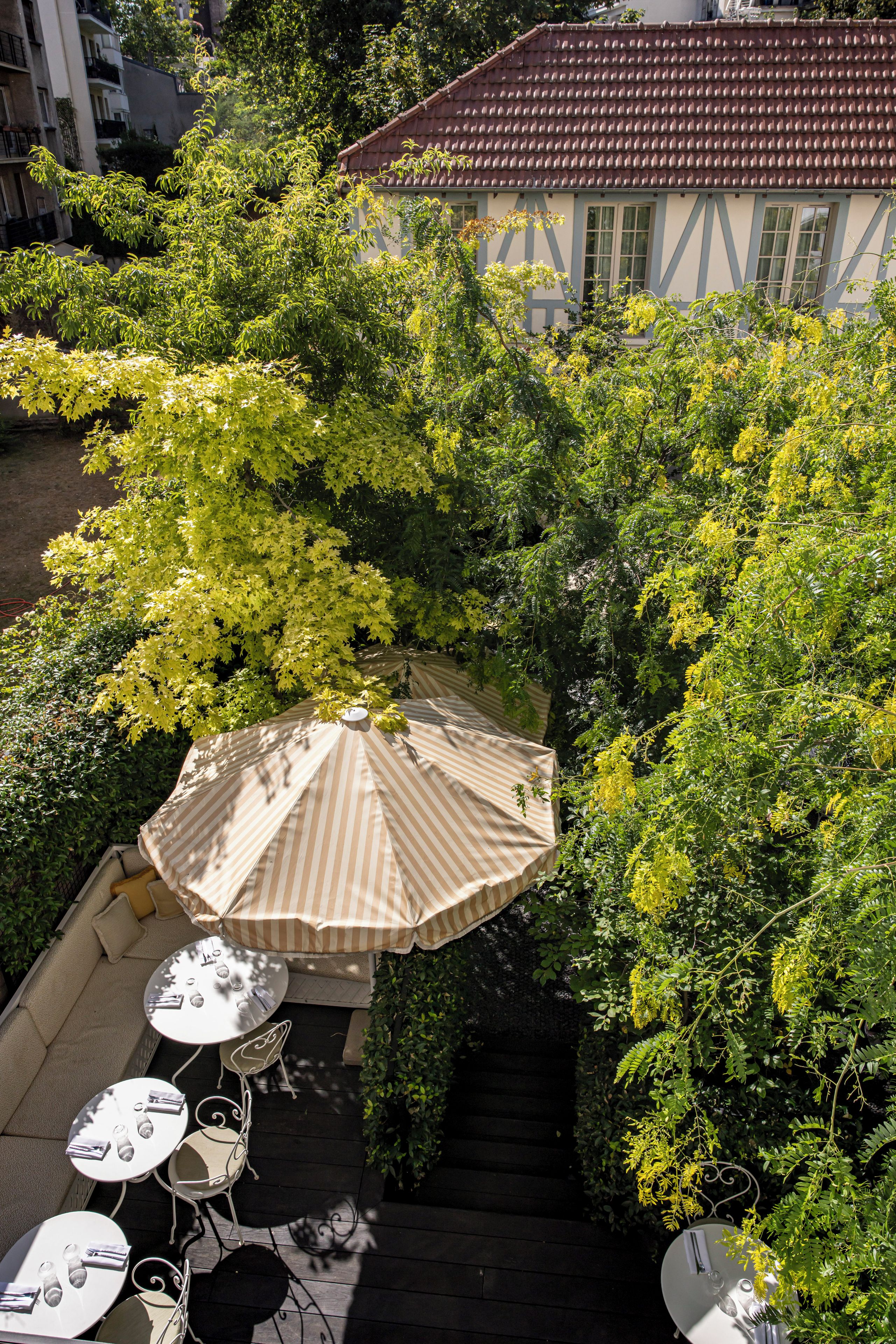 Un área de estar al aire libre con una sombrilla a rayas, mesas y sillas, rodeada de abundante vegetación y un edificio con techo de tejas al fondo.