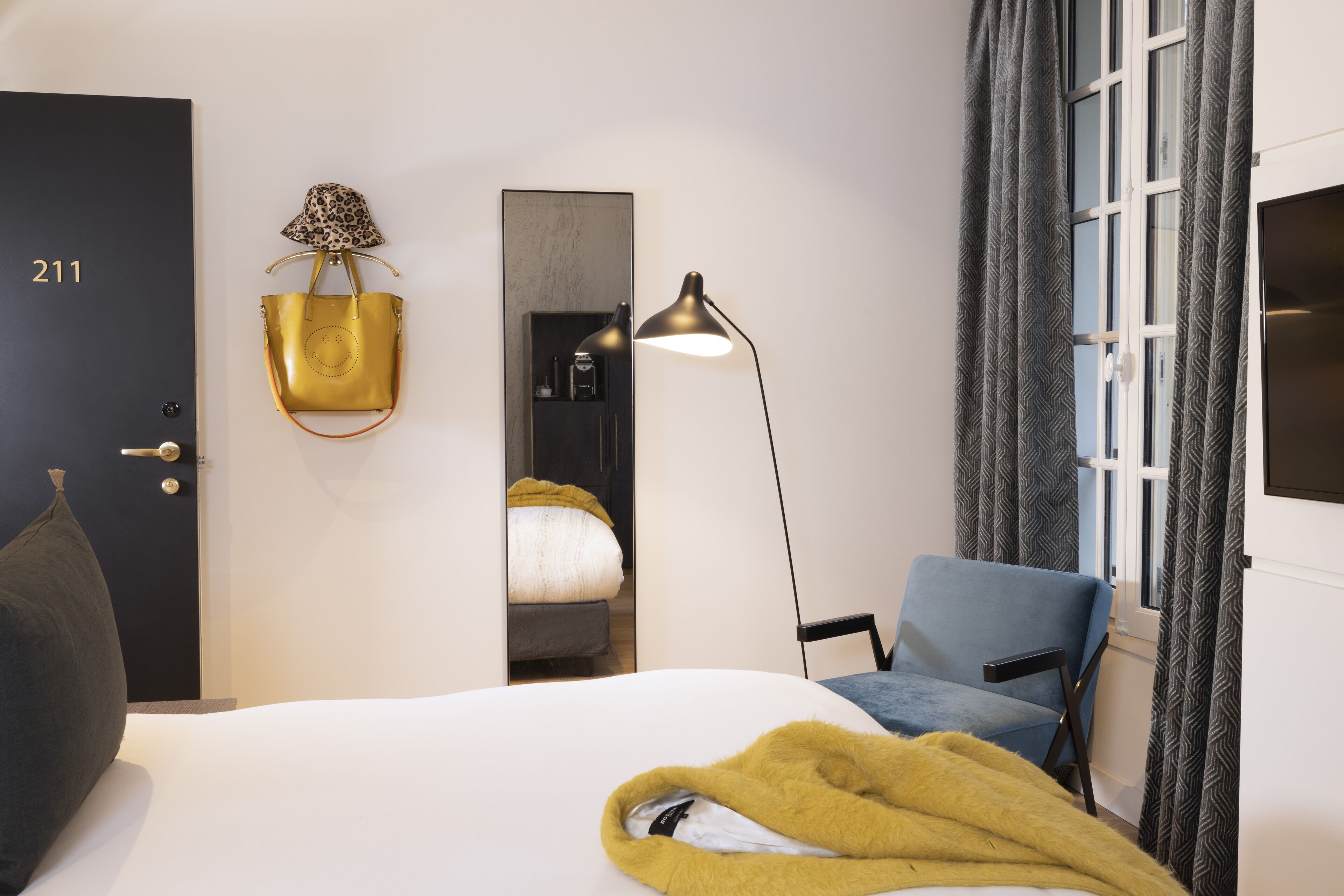 Habitación de hotel con cama, una silla, escritorio, lámpara, armario y puerta con el número 211.