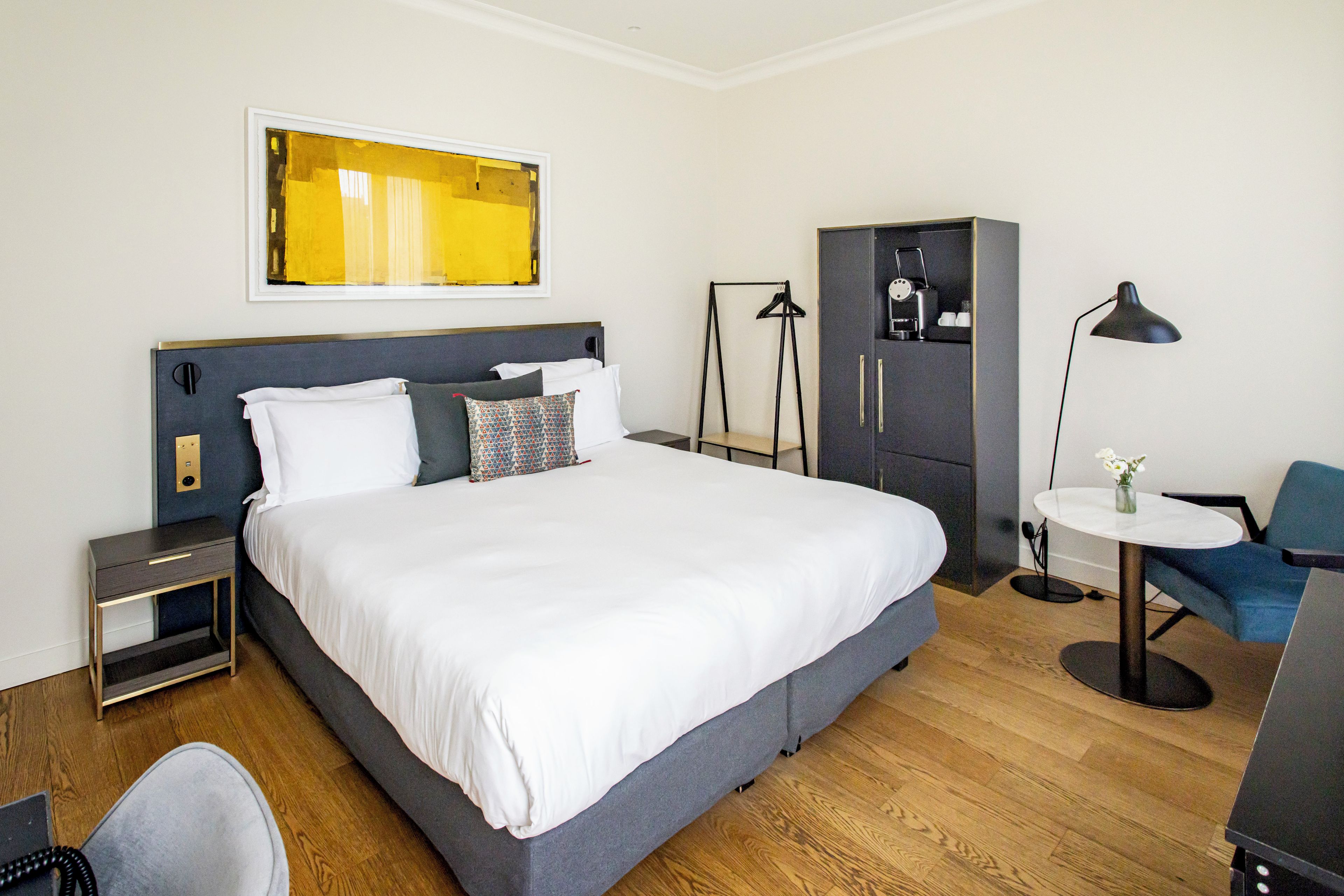 A hotel room with a large bed, a desk, a chair, a wardrobe, and a bedside table.