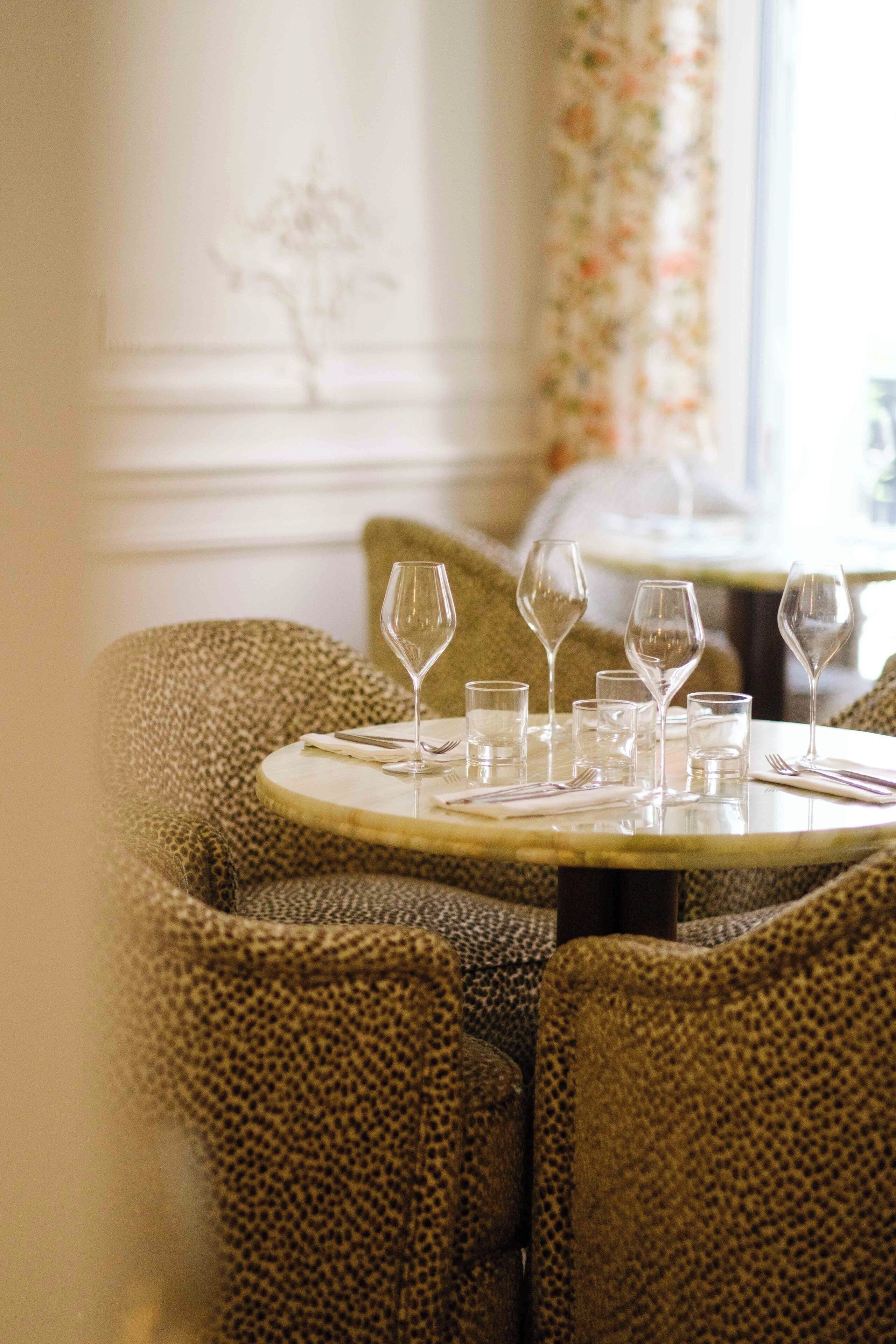 A dining table with chairs, set with wine glasses and cutlery, in a room with floral curtains and a decorative wall.