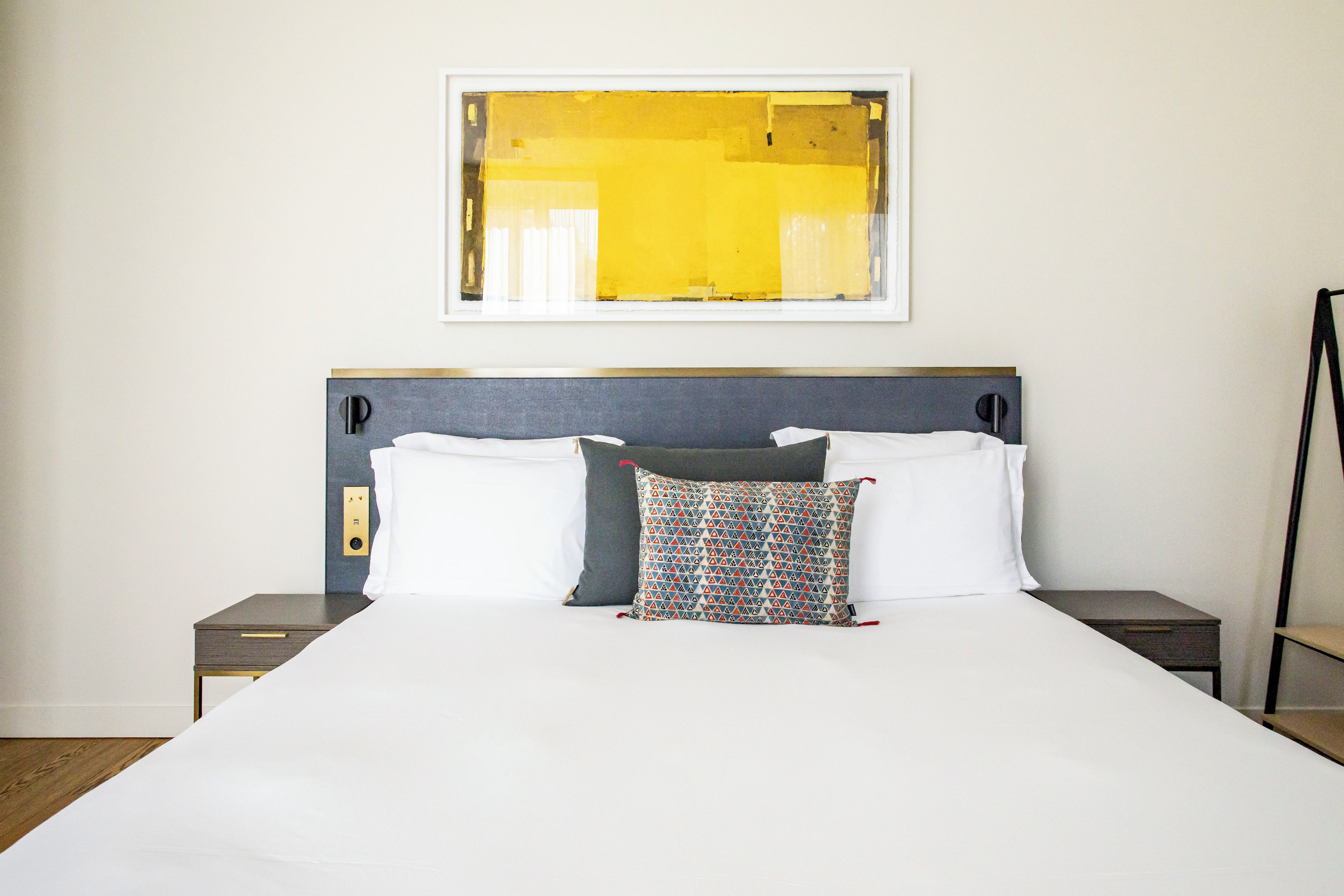 Habitación de hotel con una cama grande, una mesita de noche, un interruptor de luz en la pared y una obra de arte enmarcada sobre la cama.
