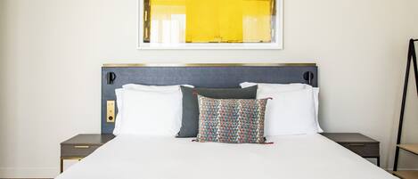 Habitación de hotel con una cama grande, una mesita de noche, un interruptor de luz en la pared y una obra de arte enmarcada sobre la cama.