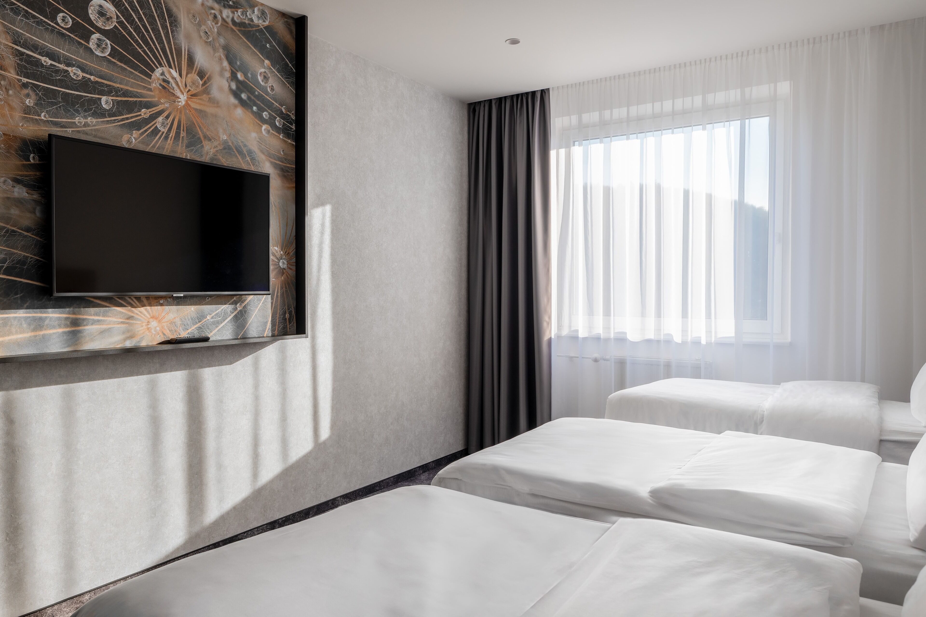 Hotelkamer met een flatscreen televisie aan een gestructureerde muur, twee eenpersoonsbedden, een groot raam met dunne gordijnen en uitzicht op de stad.