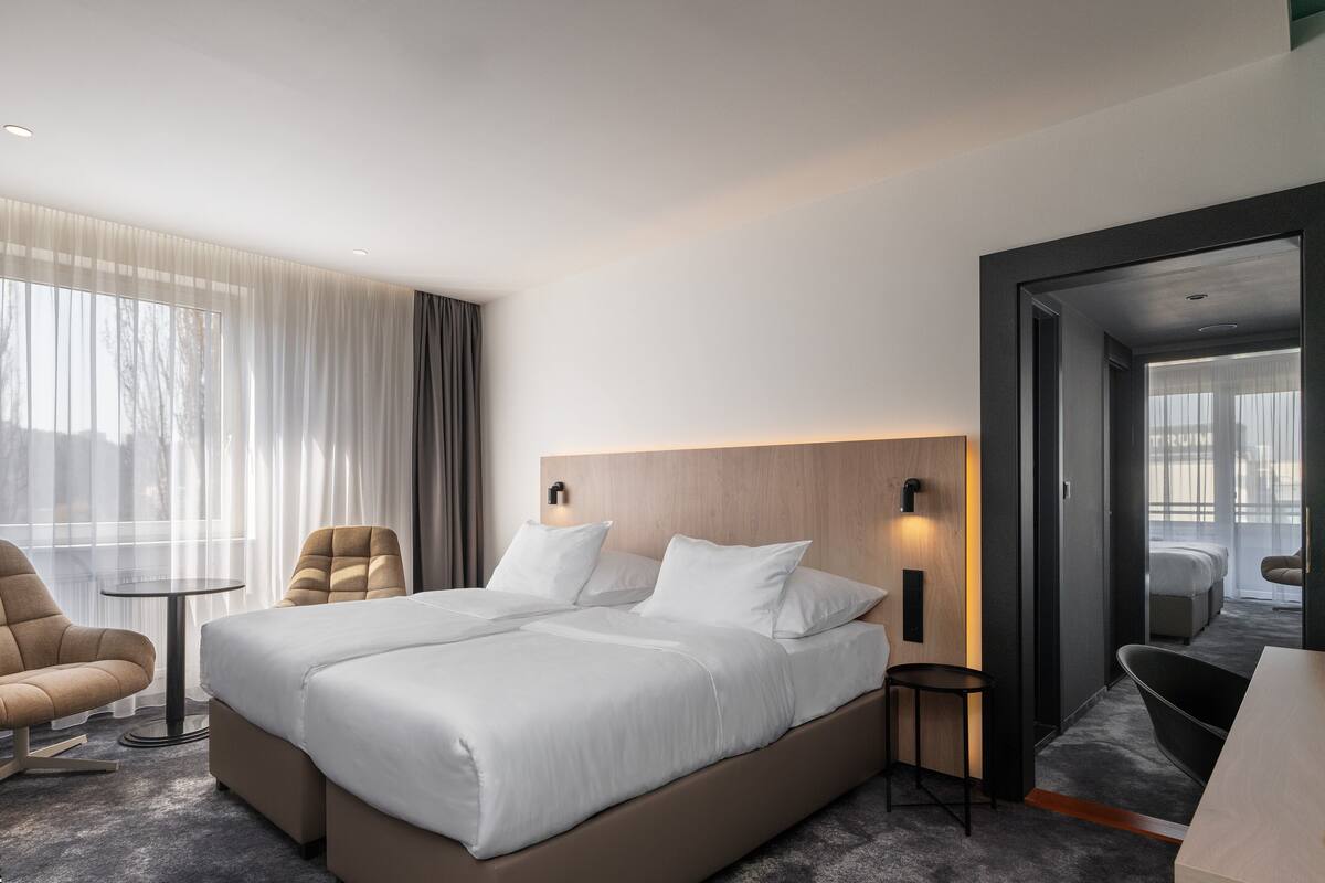 A modern hotel room with a large bed, a small table, two chairs, and a view of the city through large windows.
