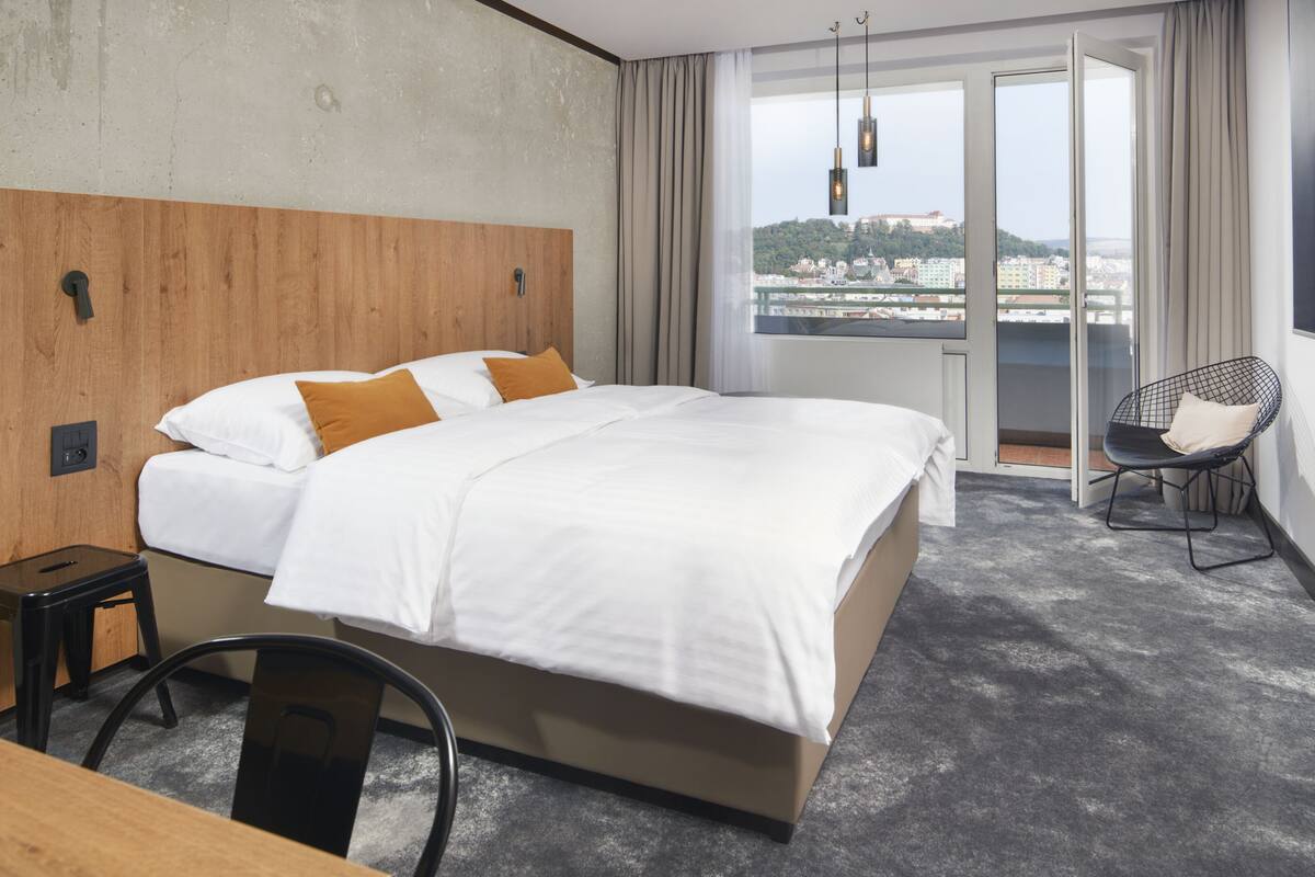 A hotel room with a bed, a chair, a desk, and a view of a cityscape.