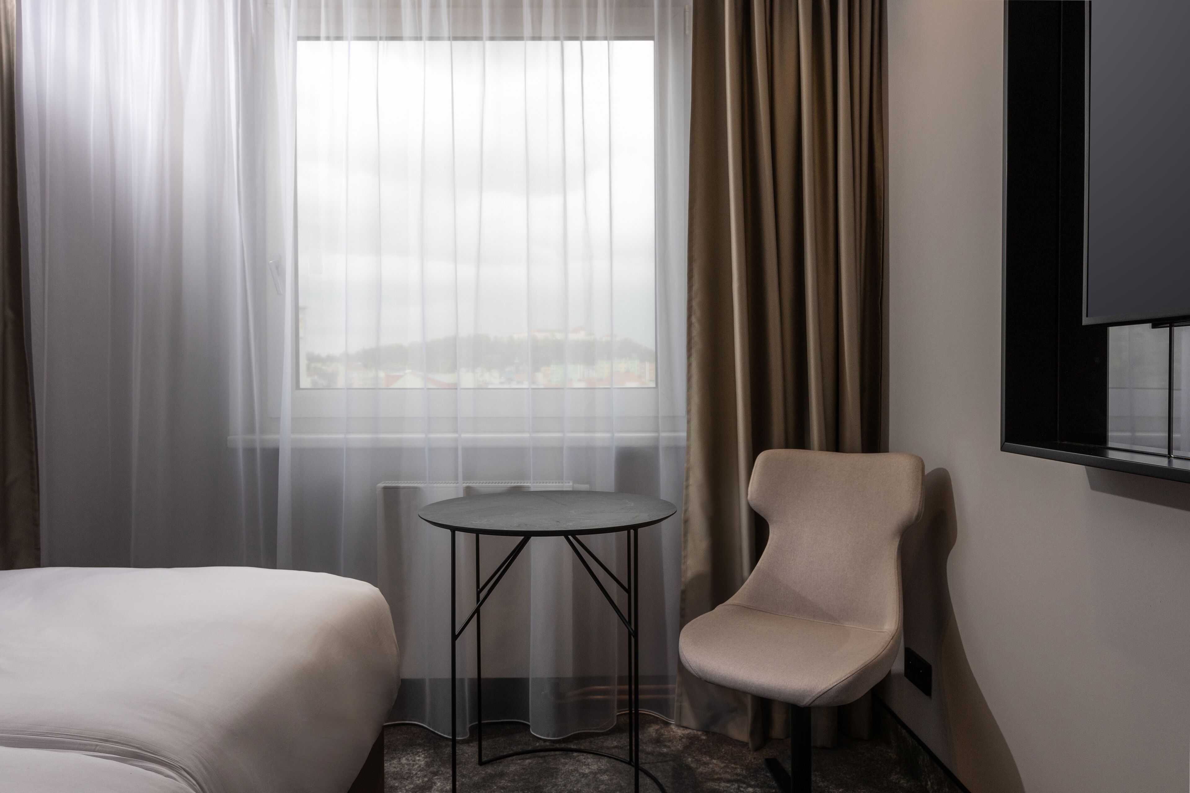 Hotelkamer met een bed, een klein rond tafeltje, een stoel, een raam met dunne gordijnen en een aan de muur bevestigde televisie.