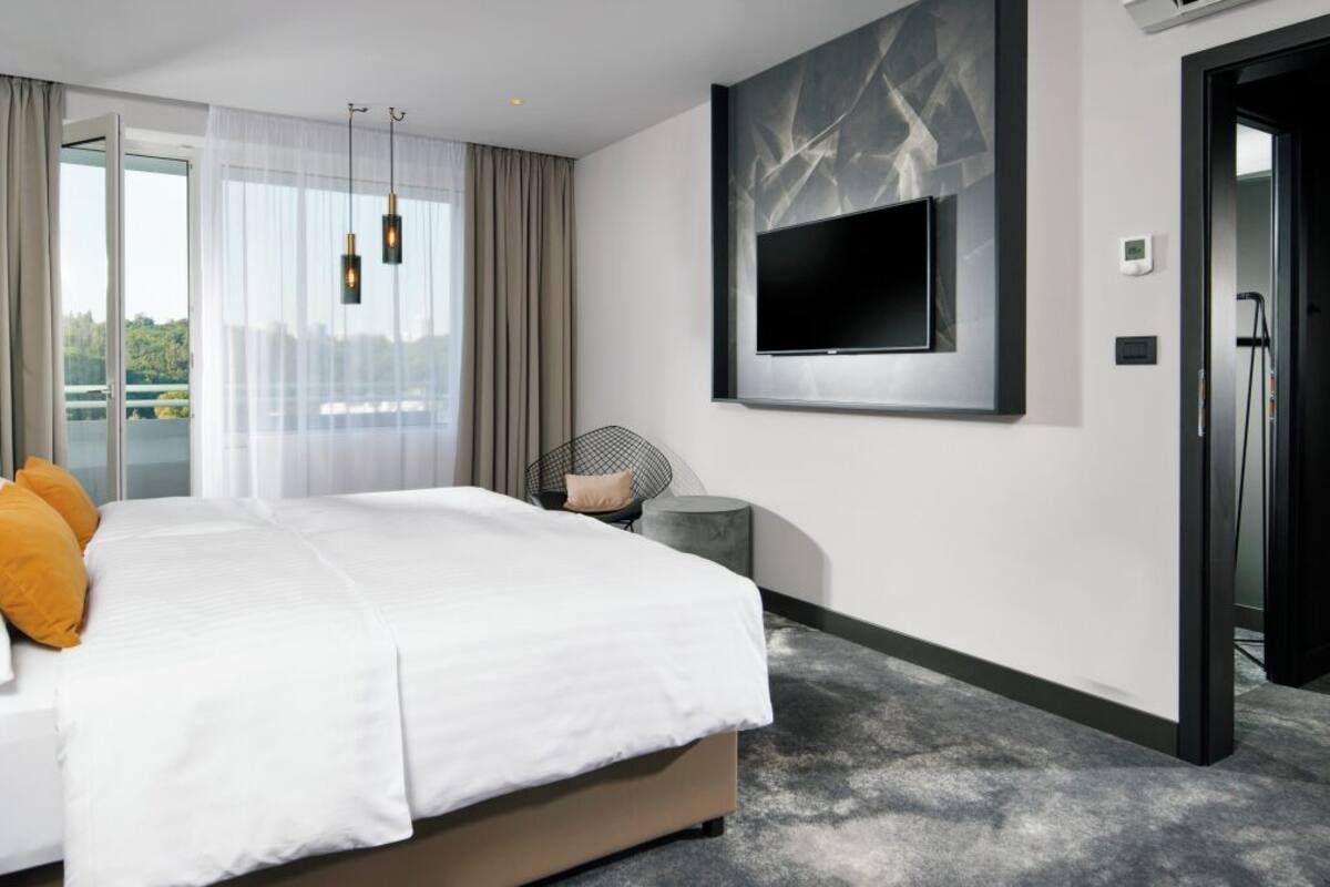 A modern hotel room with a large bed, a flat-screen TV mounted on the wall, and a sliding glass door leading to a balcony.