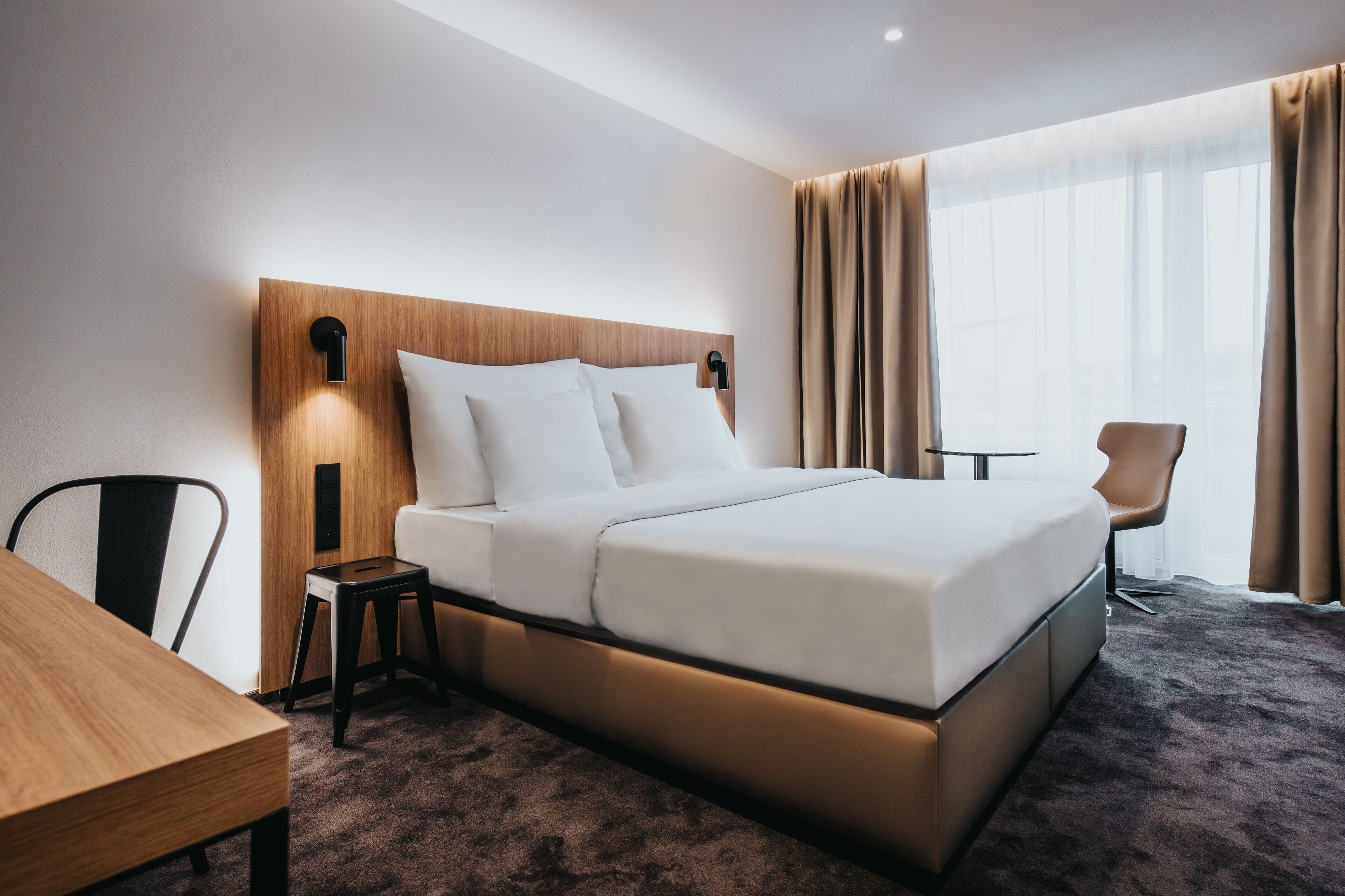 A modern hotel room with a large bed, a wooden headboard, a bedside table, a chair, and a view of the city through sheer curtains.