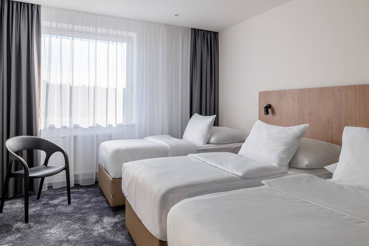 A hotel room with two beds, a chair, and a large window with sheer curtains.