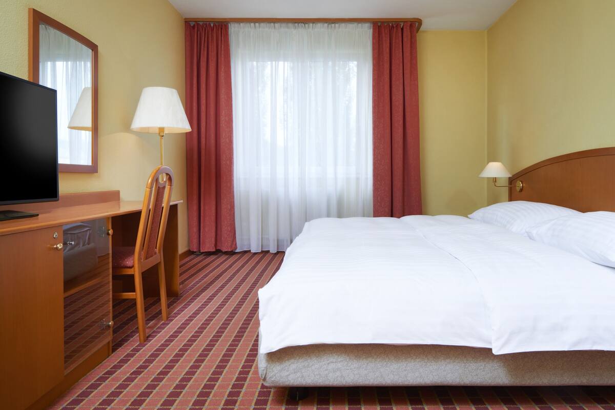 A hotel room with a bed, a desk with a television, a chair, a lamp, and a window with curtains.