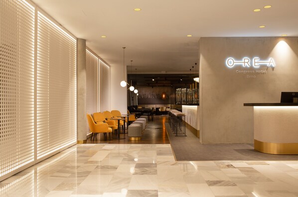 A modern hotel lobby with a reception desk, seating area, and a large window with blinds.