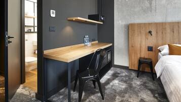 Una habitación de hotel moderna con un escritorio de madera, una silla negra, una cama con almohadas blancas y mostaza, y una pared de concreto decorativa.