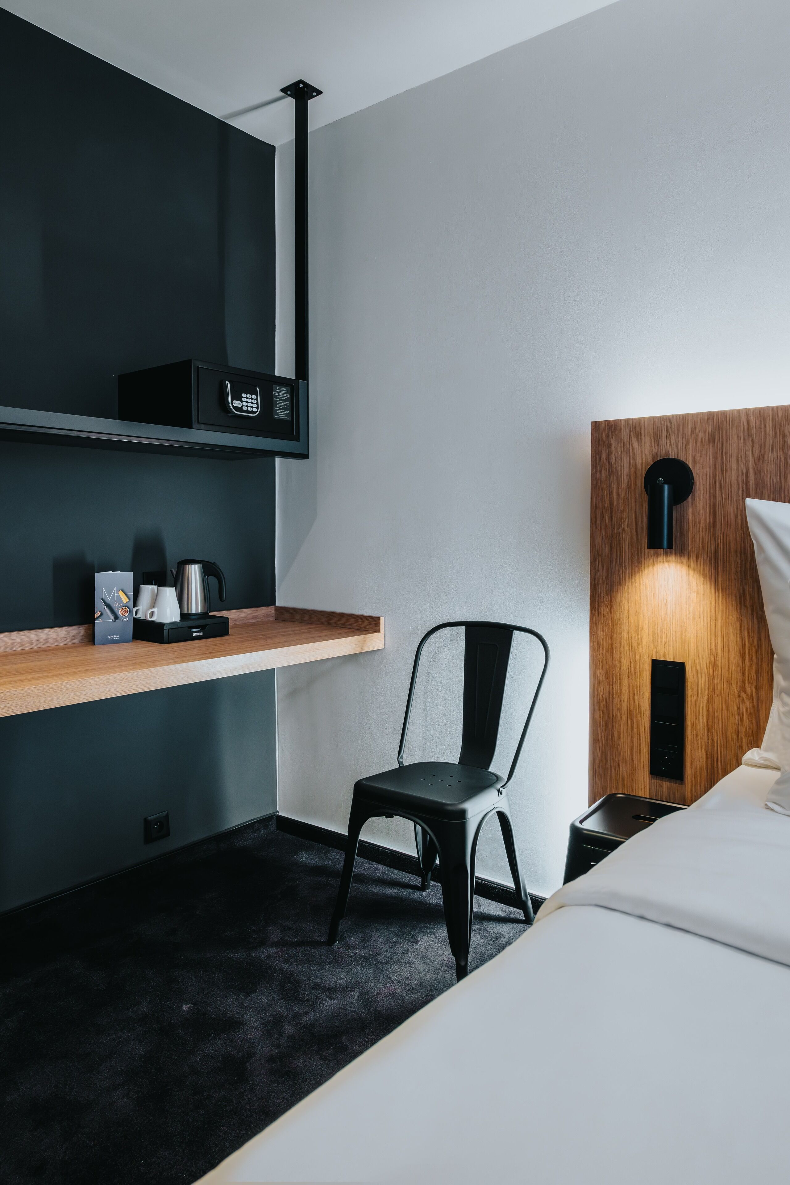 A modern hotel room with a wooden shelf, a black chair, a bed, and a wall-mounted light fixture.