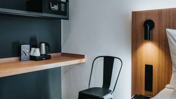A modern hotel room with a wooden shelf, a black chair, a bed, and a wall-mounted light fixture.