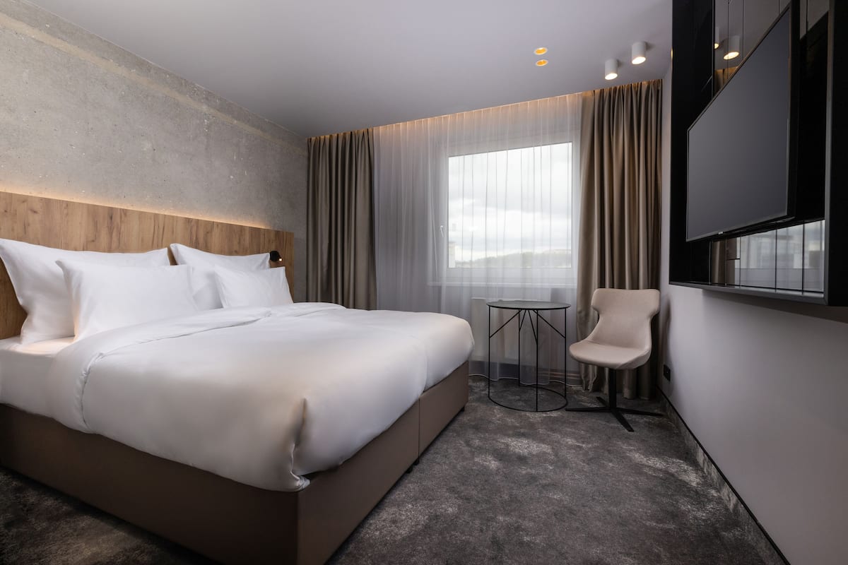 A modern hotel room with a large bed, a small round table, a chair, and a TV mounted on the wall.