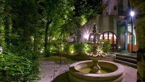Un jardín exterior nocturno con una fuente central, rodeado de árboles y un edificio con ventanas iluminadas al fondo.