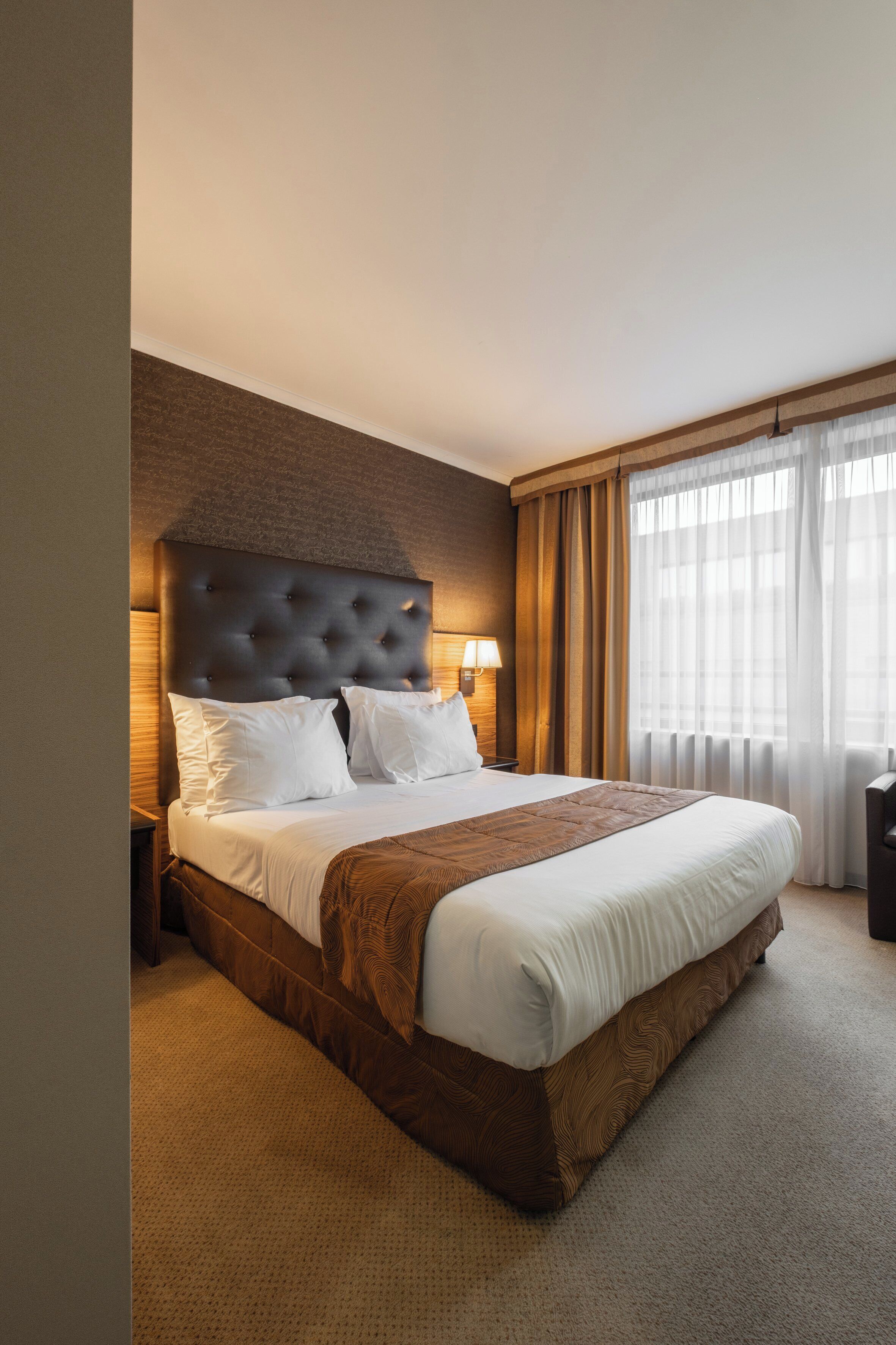 A hotel room with a large bed, a wooden headboard, a nightstand with a lamp, and a view of the outside through sheer curtains.
