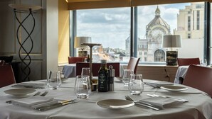 A dining table set for four with a bottle of extra virgin olive oil, glasses, and cutlery. A large window offers a view of a historic building. - Hyllit Hotel (Antwerp)