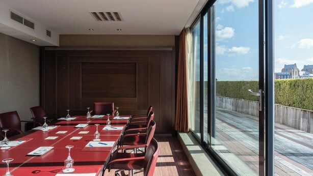 Une salle de conférence avec une longue table, des chaises rouges et une vue sur le paysage urbain, visible à travers de grandes fenêtres.