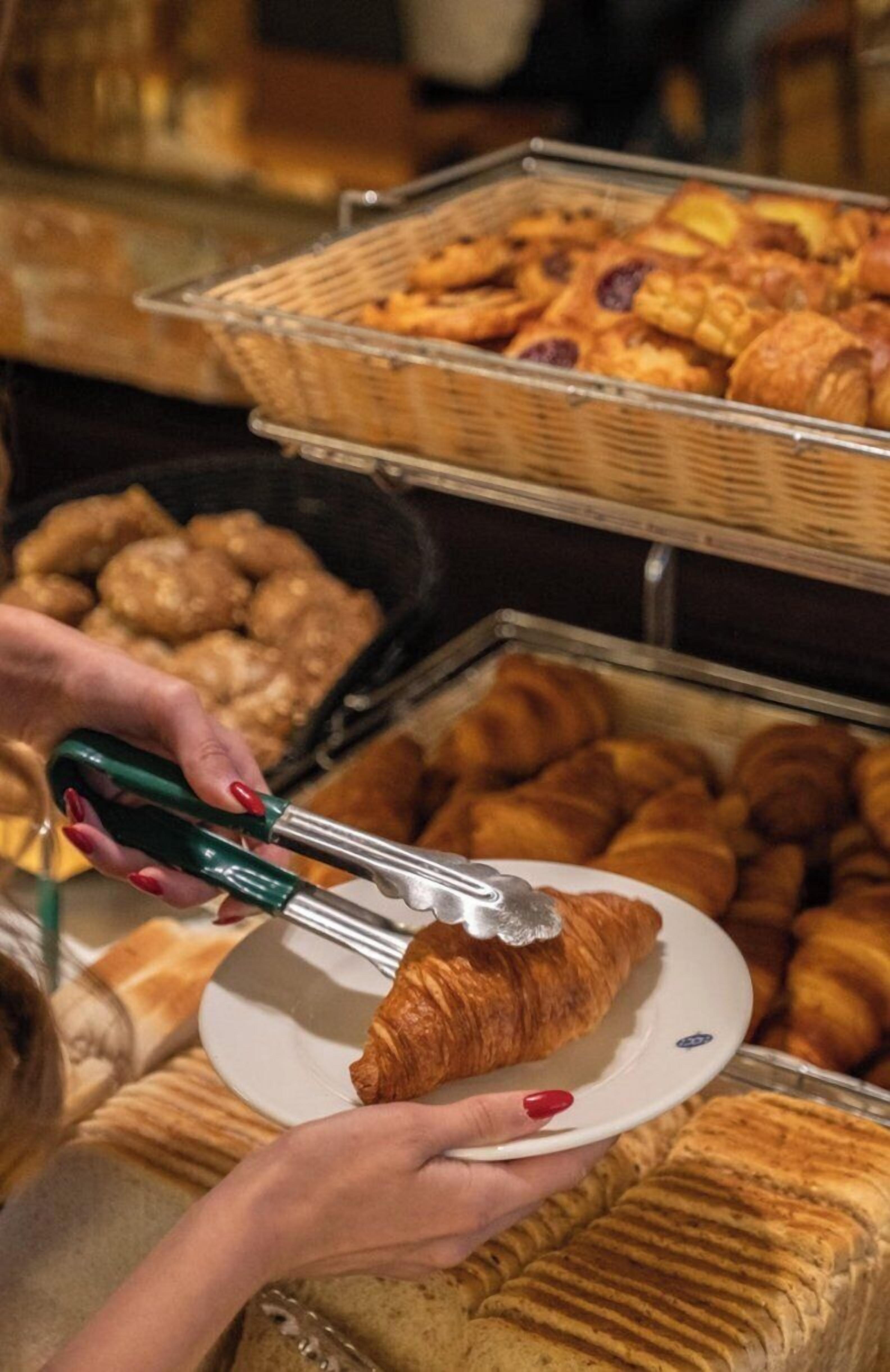 A person holding a croissant with tongs, with baskets of pastries in the background.